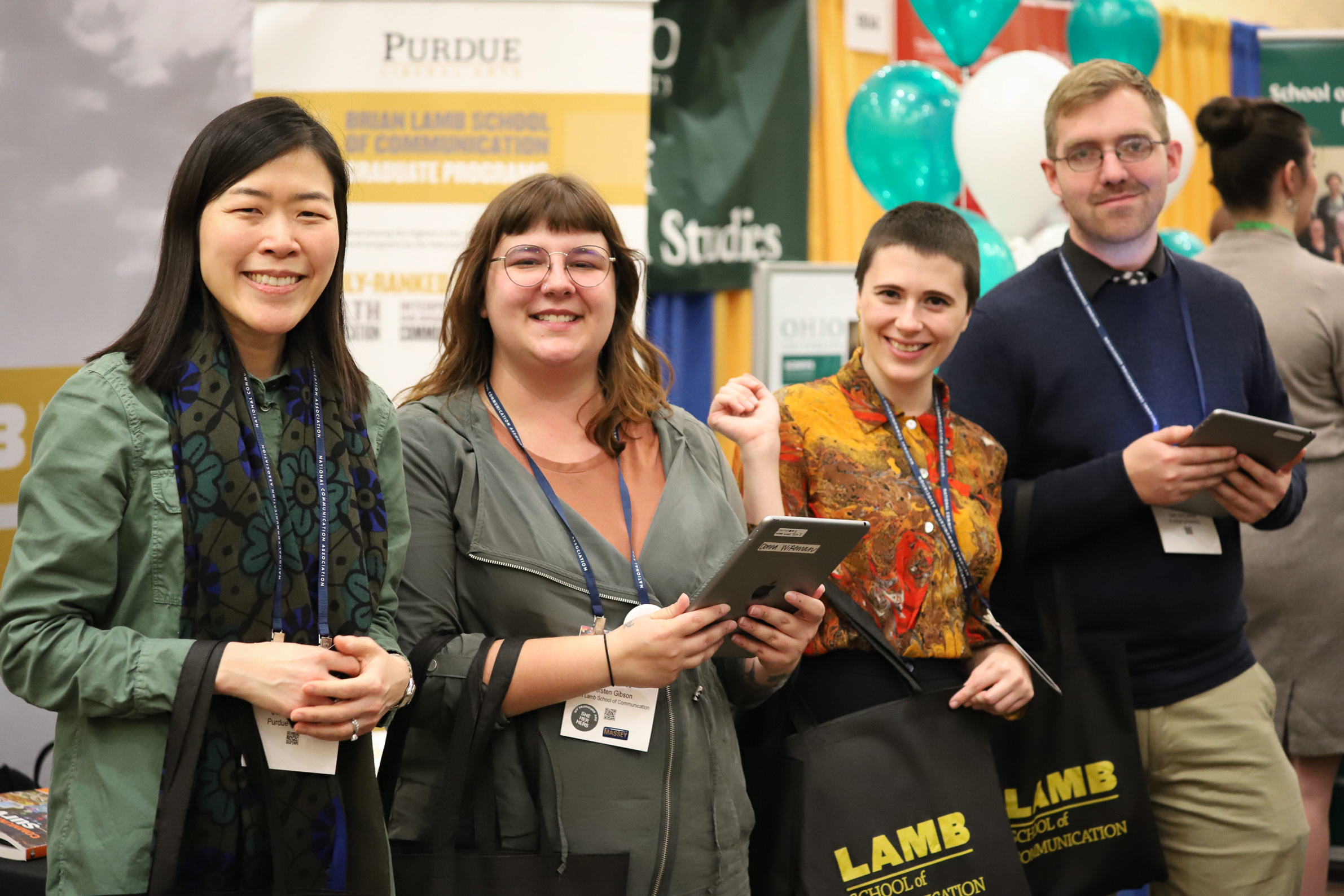 A professor and three graduate students stand together at a recent graduate recruitment fair in front of the Purdue table. They're smiling to camera. 
