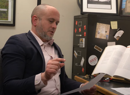 In an image taken from a video interview, Dr. Michael Johnston sits in front of a desk. He is reading from a piece of paper containing a Chat GPT article.