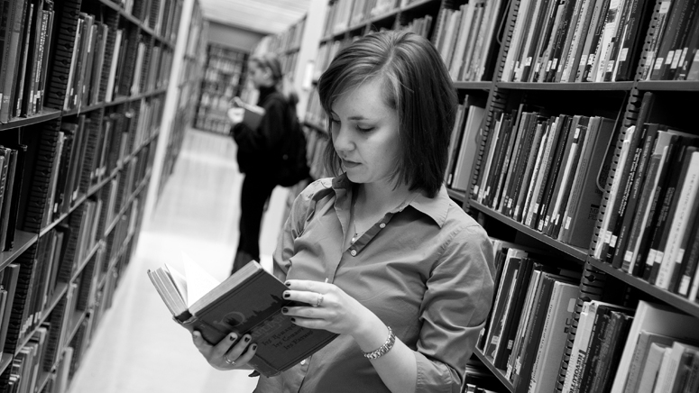 A student standing in a book aisle at a library, reading a book.
