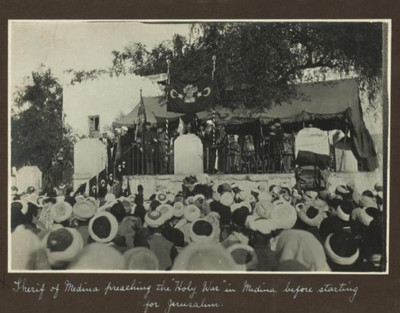 A black-and-white historical photograph with a handwritten caption stating, "Sherif of Medina preaching the Holy War in Medina before starting for Jerusalem." It shows a large crowd gathered in front of a raised outdoor platform where figures are standing under a draped canopy.