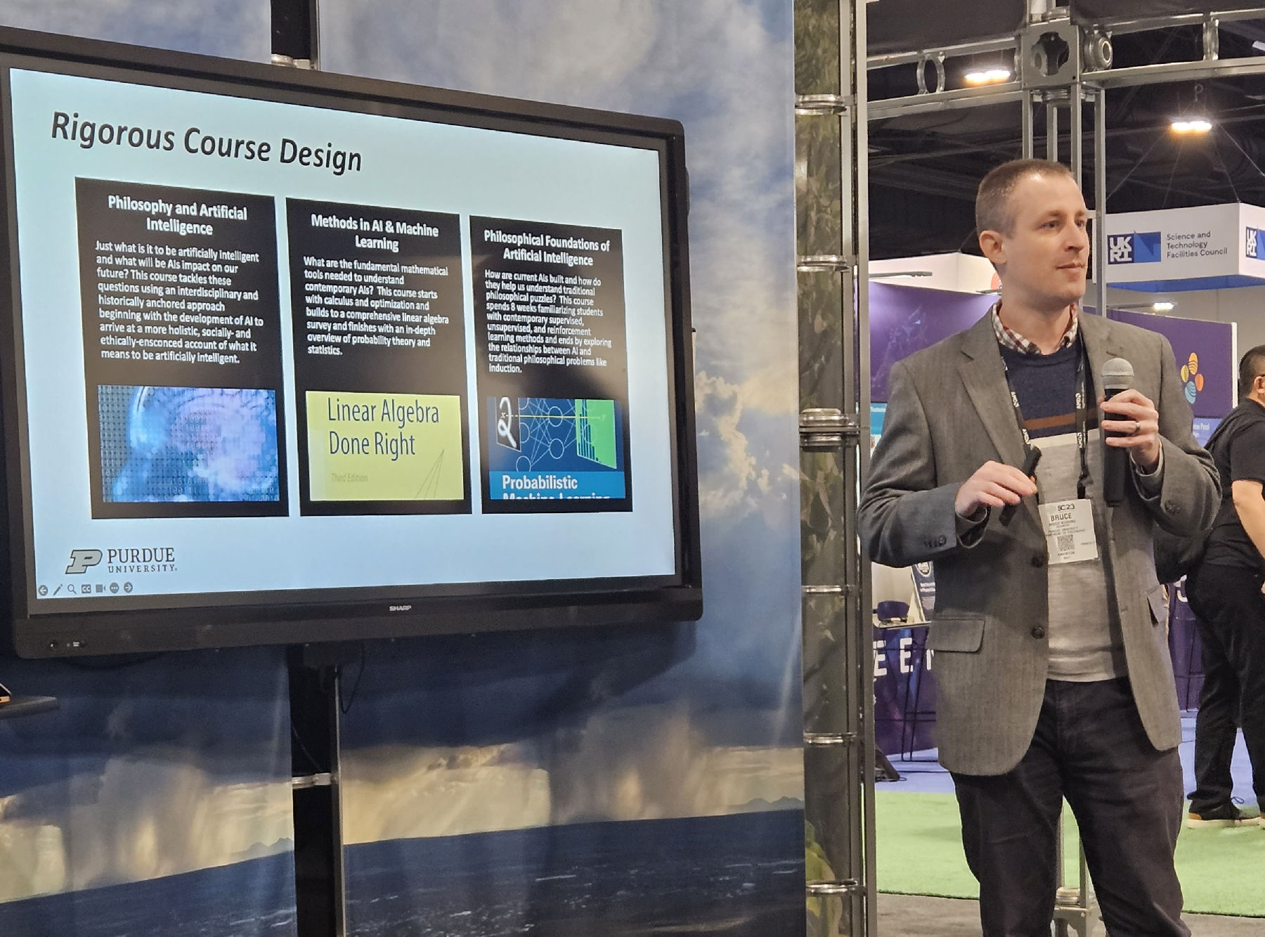 A person with a microphone, standing in front of their presentation at a convention.