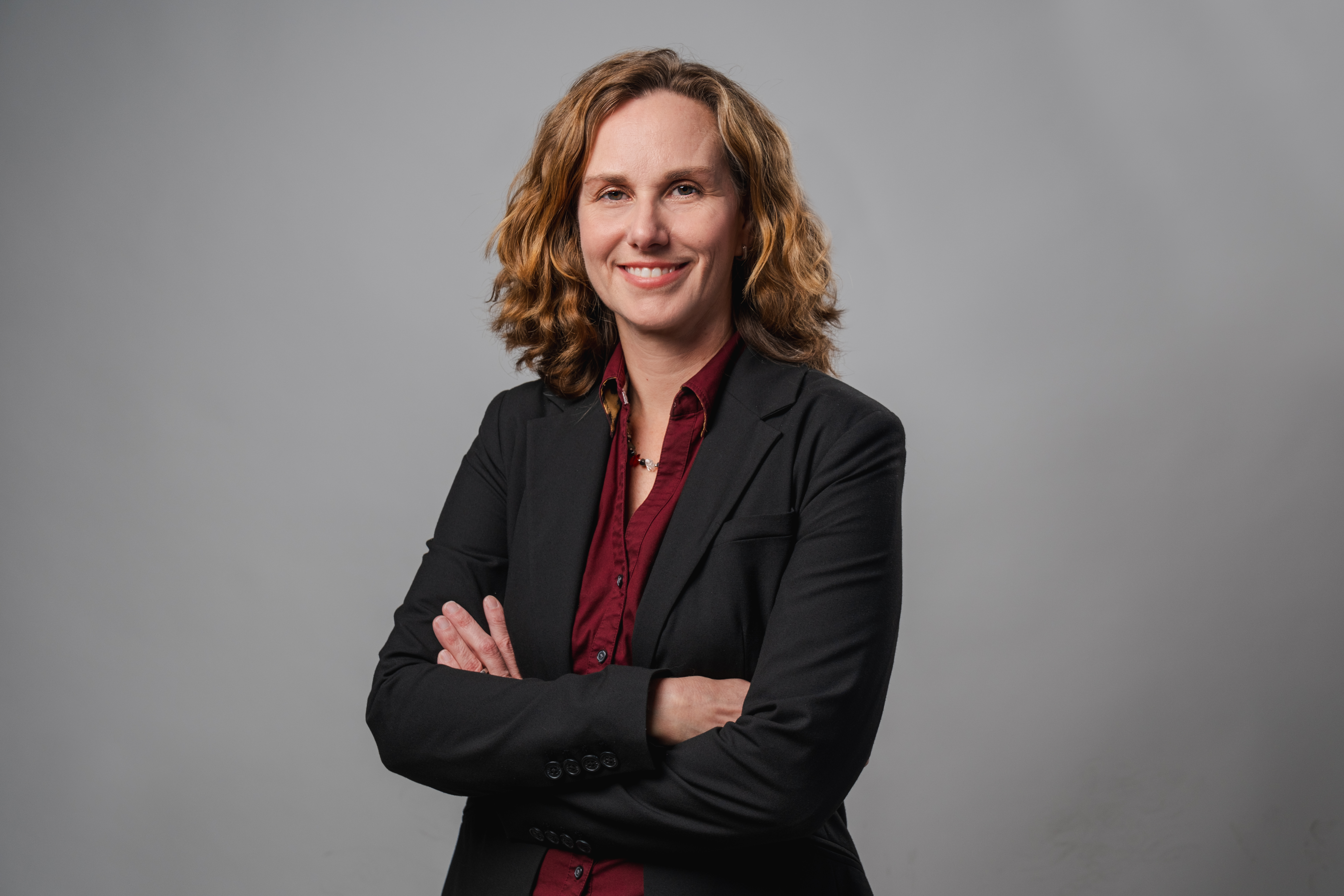 “Faculty member in a dark blazer and collared shirt standing with arms crossed against a gray background.”