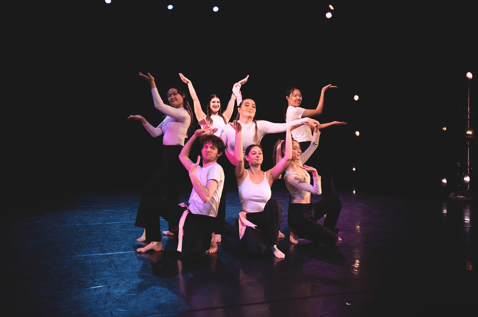 Group of dancers in staged in a pose on the floor and standing. Dancers arms are posted at various angles and heights.