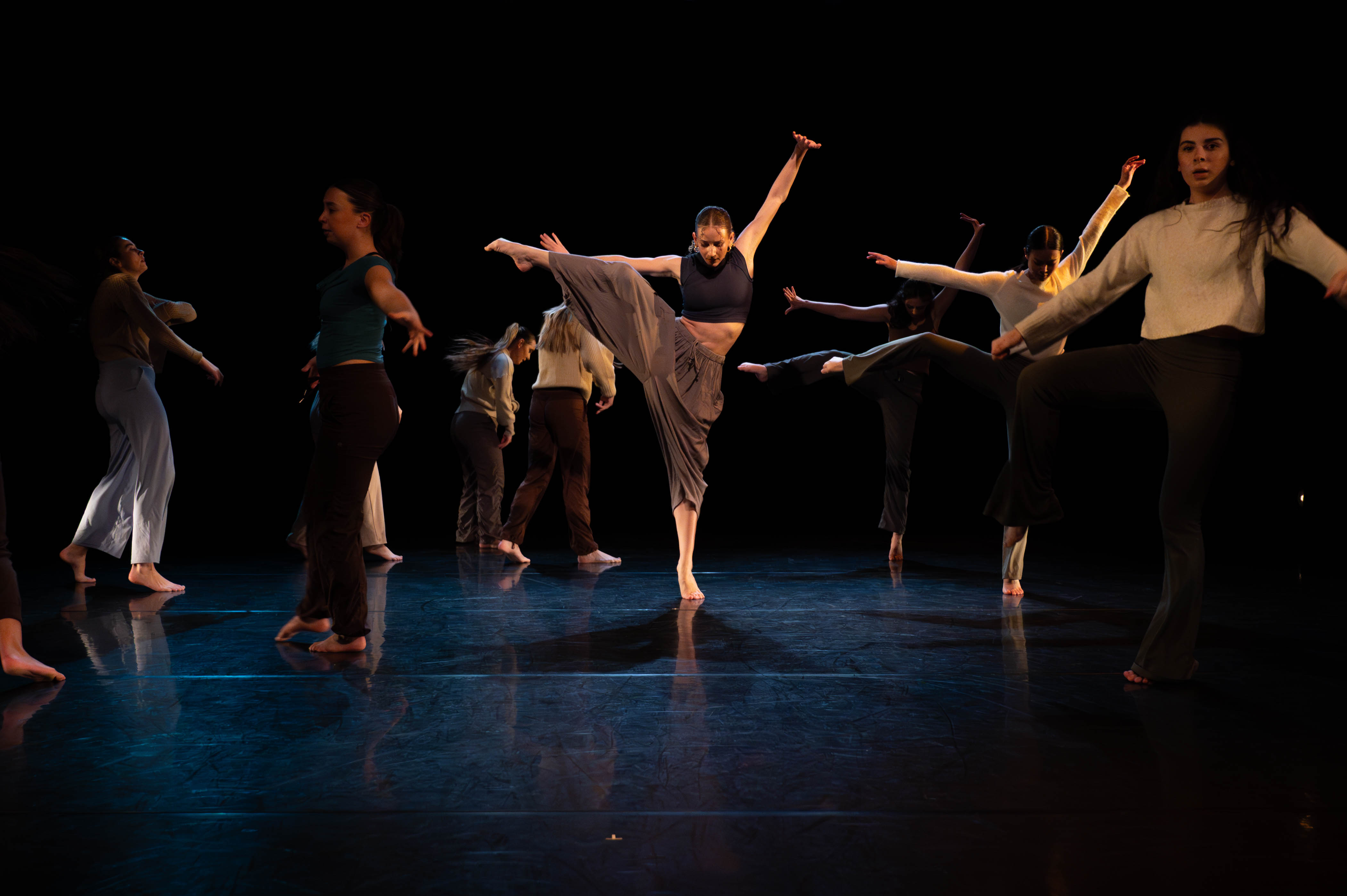 Large group of dancers on stage with one dancer highlighted in the center in. Dancer is in a pose with one leg extended out to the side and arms in the air.