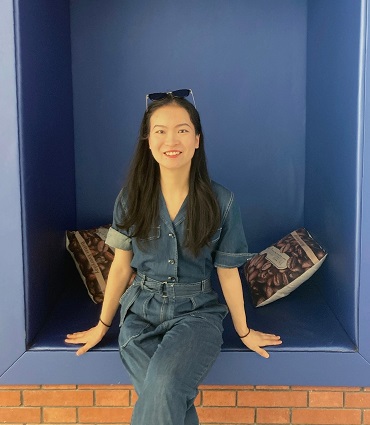 A photo of Yue Li. A woman with black hair, black eyes, and sunglasses atop her head sitting and smiling in a denim romper in an alcove.