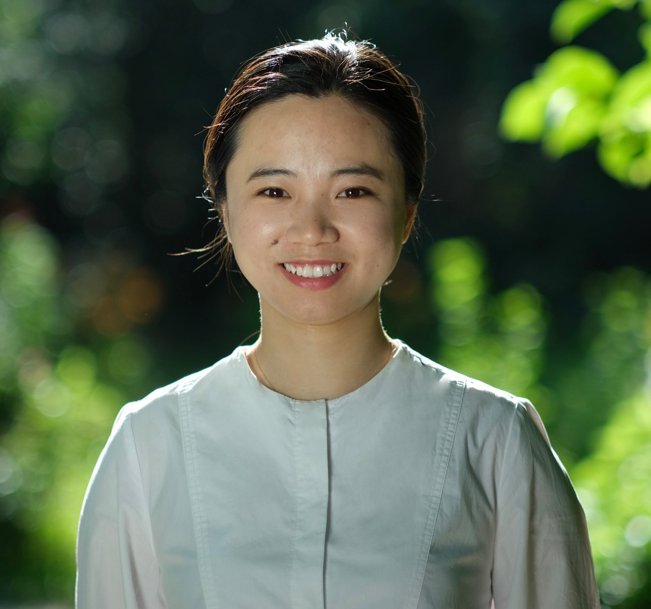A photo of Jingying Hu. A woman with black hair, black eyes, and a white top smiling against a sunlit leaves in soft focus. 