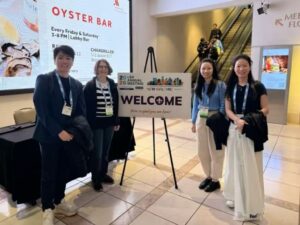 Four people dressed in academic conference clothes, standing besides a sign that says Welcome