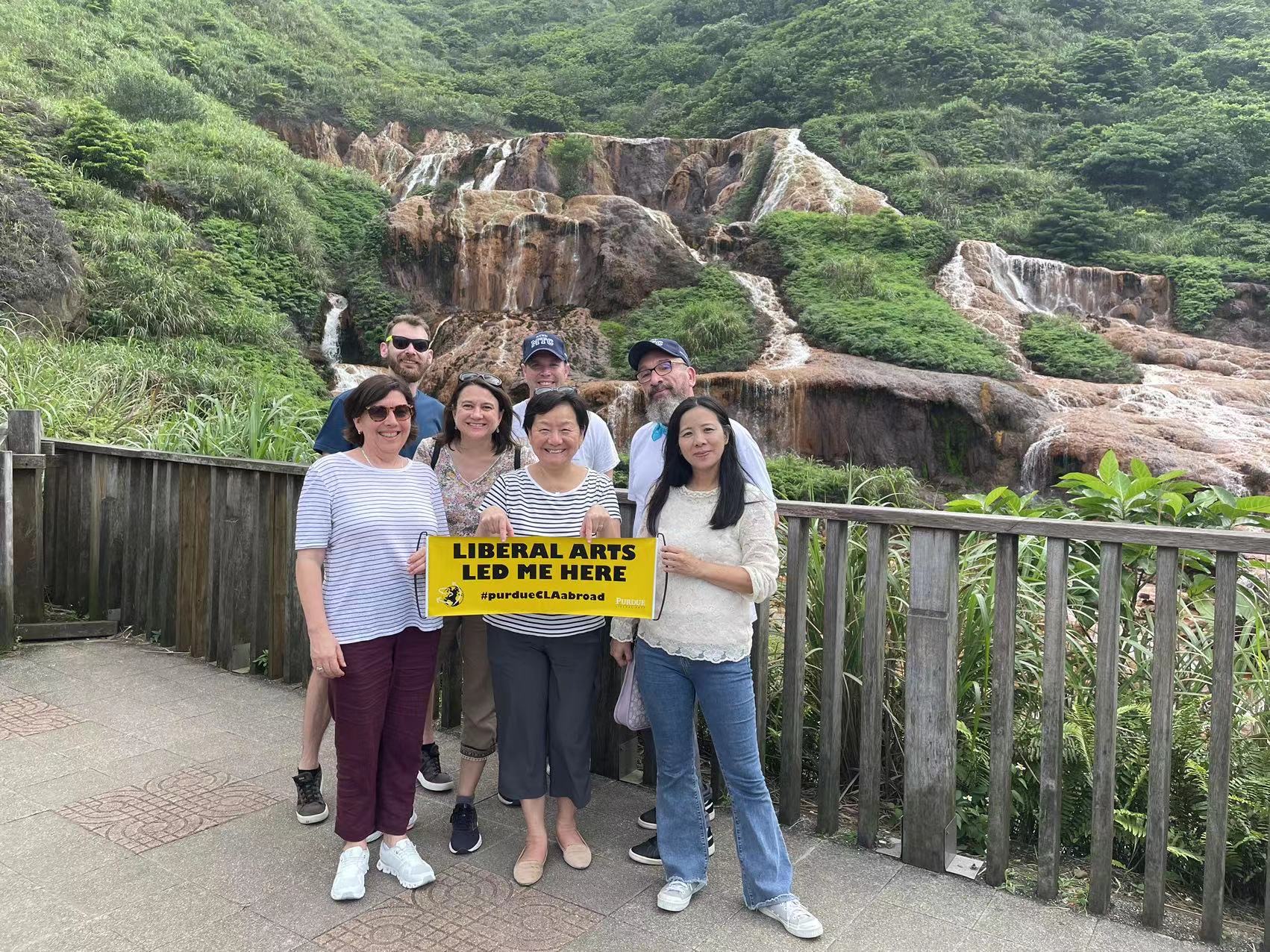 Group of CLA administrators smiling for a photo at a waterfall in Taiwan holding a "Liberal Arts led me here" sign.