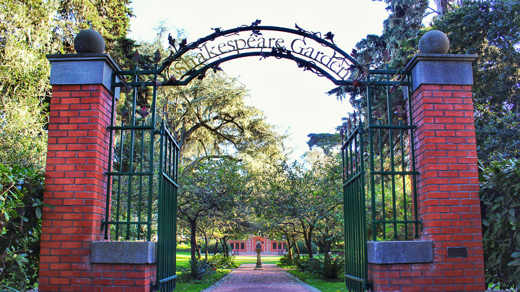 A park entrance with a brick and green iron gate with the words Shakespeare Garden on the arch above it
