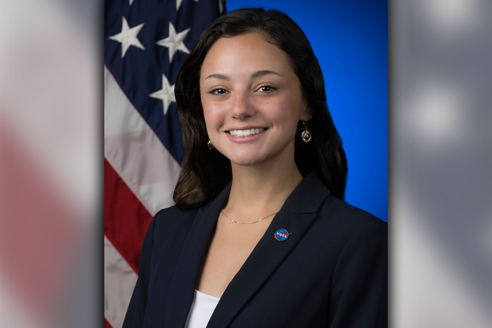 Lucia Morton smiling in front of American flag, dressed in navy blue suit with NASA pin.