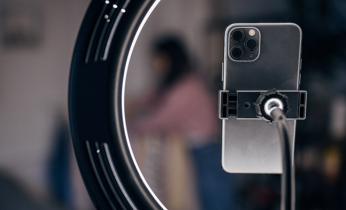 A ring light camera with an IPhone three lens camera with a woman blurred behind it.