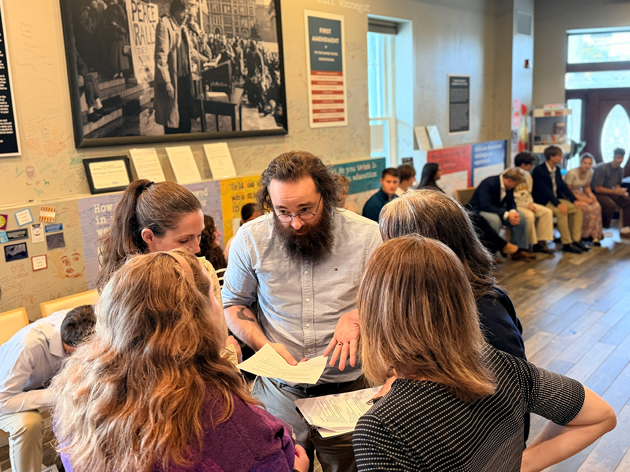 Group of faculty stand in a circle holding papers talking in a room full of students.